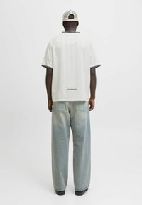 Hombre con camisa blanca de manga corta, jeans anchos de color azul claro, zapatos negros y gorra de malla beige, de pie de espaldas sobre un fondo blanco liso.