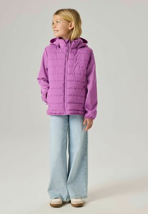 Copil cu păr blond, purtând o jachetă matlasată mov cu glugă, blugi largi de culoare albastru deschis și pantofi albi, uitându-se într-o parte și zâmbind.