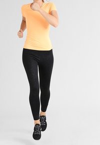 Lyhyet hihat omaava tiukasti istuva oranssi urheilu t-paita on yhdistetty mustiin kuviollisiin leggingseihin. Varustukseen kuuluvat nauhalliset urheilukengät.