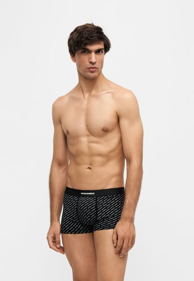 Jeune homme aux cheveux foncés portant des boxers noirs à motifs, debout devant un fond clair uni.