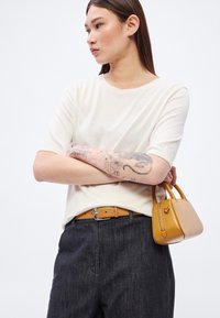 Svetlo bež pulover s kratkimi rokavi, temno denim hlače in rjav usnjen pas. Majhna rumena torbica z zadrgo v roki.