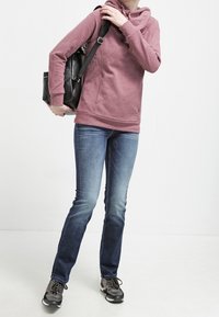 Felpa leggera color marrone con tasca frontale e cordini, abbinata a jeans blu e scarpe da ginnastica nere, mentre porta uno zaino nero.
