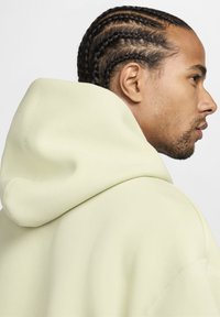 Svetlo zelena kapuca z mehko teksturo, opremljena z veliko kapuco in brezšivno zasnovo. Stranski pogled prikazuje pletene lase in obrazni profil.