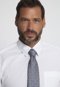 Chemise blanche à col classique, associée à une cravate bleue à motifs avec de petites fleurs, sur fond neutre.