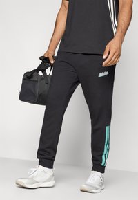 Pantaloni de sport negri cu un design ajustat, evidențiind accente teal cu trei dungi pe lateral și un logo brodat. Purtați cu încălțăminte sport.