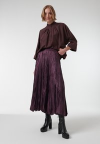 Donkerbruine, losse blouse met hoge kraag en knoopsluiting, gecombineerd met een lange, geplooide paarse rok en zwarte platformlaarzen.