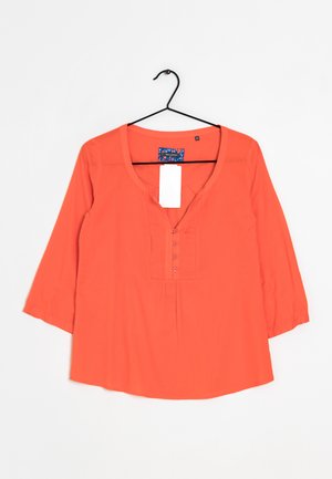 Blusa arancione brillante a maniche lunghe con scollo a V, quattro bottoni e vestibilità ampia appesa a una gruccia nera su sfondo bianco.