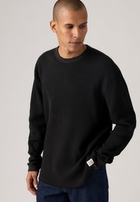 Black long-sleeve ribbed sweater with a round neckline, textured fabric, and a visible brand tag at the hem. Worn with blue jeans.