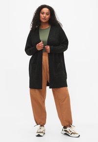 Schwarze Cardigan mit offenem Vorderteil und zwei Taschen, getragen über einem grünen Oberteil und bequemen, beige-farbenen Pants. Das Schuhwerk hat ein mehrfarbiges Design.