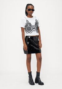 Balta trumpa marškinėliai su juoda nėriniu, derinami su juoda blizgučių sijonu. Modelis mūvi juodus akinius nuo saulės ir masyvius juodus batus.