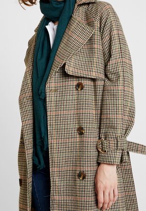 Femme portant un trench-coat à carreaux marron, beige et vert avec des boutons en écaille, une écharpe bleu sarcelle foncé, un haut blanc et un jean bleu foncé.