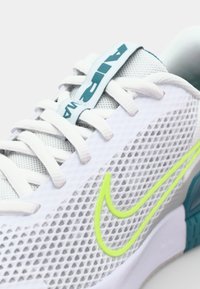 White mesh athletic shoe with light teal accents, yellow swoosh logo, and flat laces. Features ventilated upper and cushioned sole.