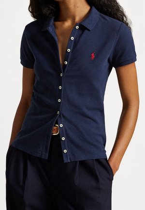 Vrouw draagt een marineblauw kort mouw poloshirt met knopen en een rood geborduurd logo op de linkerborst, ingestopt in een donkere broek.