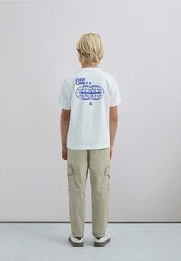 Camiseta blanca de algodón con un estampado gráfico azul en la parte posterior que dice "FUERA DE LÍMITES" y un diseño de globo, combinada con pantalones cargo beige.