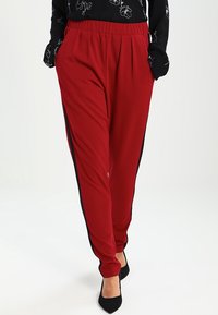 Rode joggingsbroek met zwarte zijstrepen, gemaakt van zachte stof, met plooien aan de voorkant en een elastische tailleband. Zwarte hoge hakken.