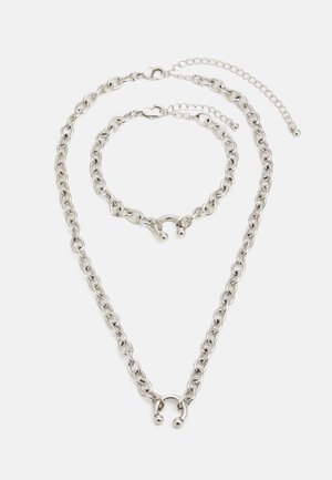 Srebrna metalna verižica in zapestnica. Debele povezave s poliranim zaključkom, opremljene z zapiranjem in prilagodljivo dolžino. Enostaven dizajn.