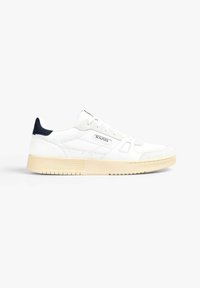 Zapatilla blanca con un cuerpo texturizado, acentos de gamuza y suela de goma beige. Presenta un talón en navy y una marca en el costado.