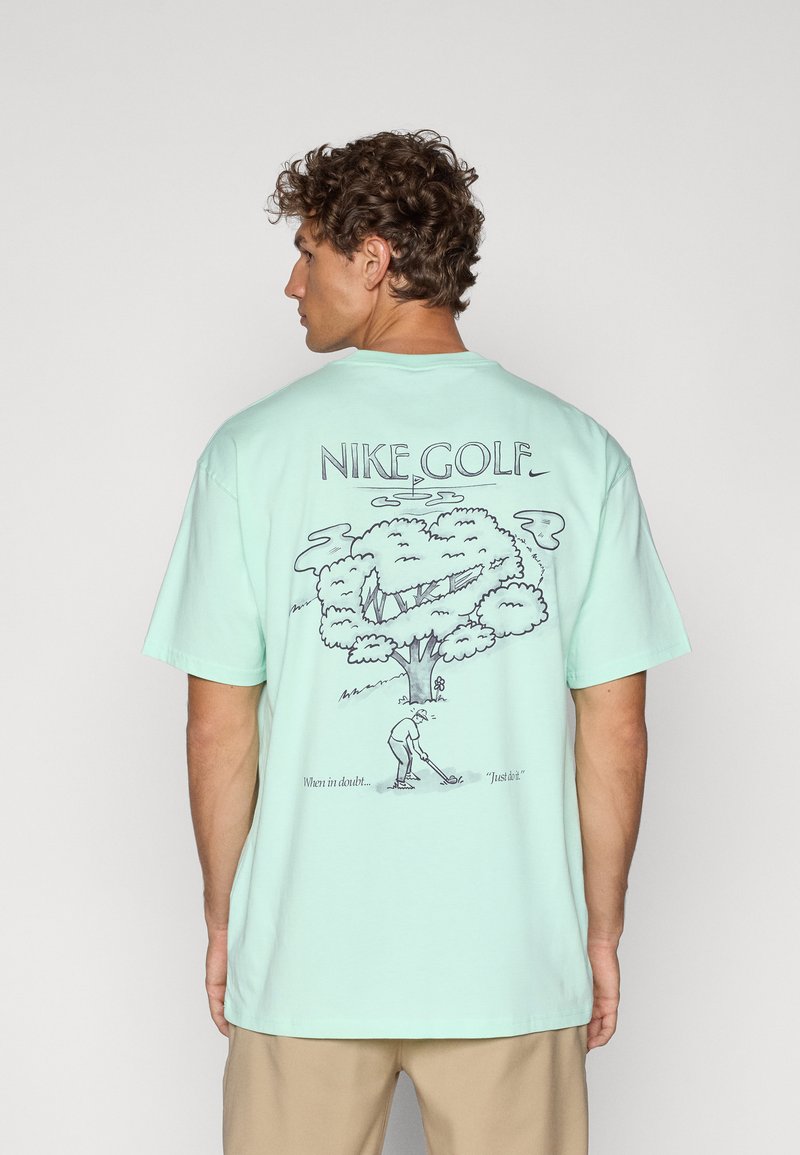 Mintgrøn bomulds t-shirt med et grafisk print af et træ og en golfspiller på ryggen, med teksten "Nike Golf" og sætninger nedenfor illustrationen.