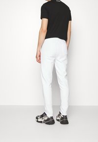 Bílé slim fit tepláky s kapsou na zadní straně, v kombinaci s černým tričkem a šedoběžovými teniskami. Hladká textura.