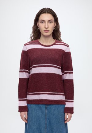 Femme aux cheveux bruns ondulés portant des boucles d'oreilles anneaux dorés, un pull rayé bordeaux et rose clair, et une jupe en jean bleue, debout devant un fond blanc.