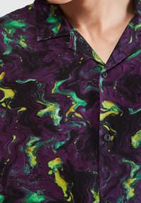 Chemise à manches courtes présentant un motif marbré en violet, vert et jaune. Col boutonné ; le tissu semble lisse avec une légère brillance.