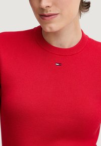 Maglione a coste rosso con scollo rotondo e piccolo logo ricamato in blu navy, bianco e rosso vicino al colletto. Morbida texture.