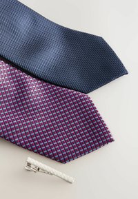 To sæt slips: et marineblåt slipset med et tekstureret gittermønster, et bourgognefarvet slipset med et geometrisk design i lilla og lyseblå, plus en sølvfarvet slipsenåle.