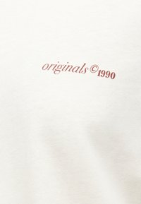 Κείμενο "originals © 1990" τυπωμένο σε καφέ χρώμα σε εκρού υφαντό φόντο.