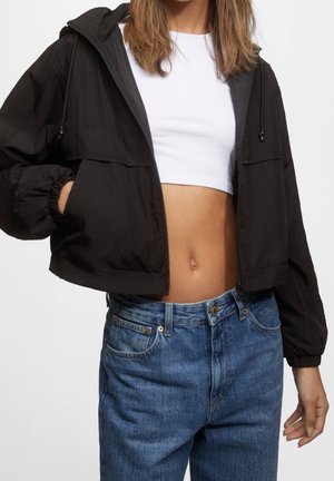 Frau trägt eine kurze schwarze Jacke über einem weißen Crop-Top und hoch taillierte blaue Jeans, mit einer Hand in der Jackentasche.