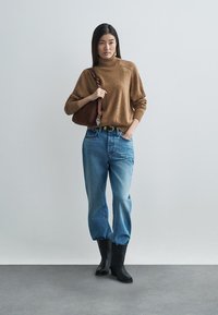 Καφέ πλεκτό πουλόβερ, μπλε τζιν με ψηλή μέση και χαλαρή εφαρμογή, μαύρα μποτάκια αστραγάλου και καφέ τσάντα· μινιμαλιστικός σχεδιασμός σε ουδέτερο φόντο.