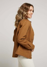 Veste marron à boutons avec un tissu texturé, poches latérales et coupe décontractée, associée à un pantalon de couleur claire.