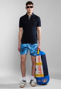 Polo shirt navy con maniche corte, collare con motivo, pantaloni da bagno blu a fantasia, e che porta una borsa da palestra multicolore. Indossa occhiali da sole.