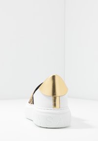 Biała trampka z teksturowaną czarną bokiem, złotym akcentem na pięcie i wyraźnym logo na grubej, białej podeszwie. Gładka skóra i połyskujące wykończenie.