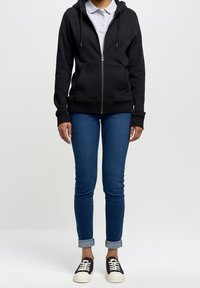 Felpa nera con zip, cappuccio e tasche frontali, abbinata a jeans denim blu slim-fit e sneakers bianche e nere.