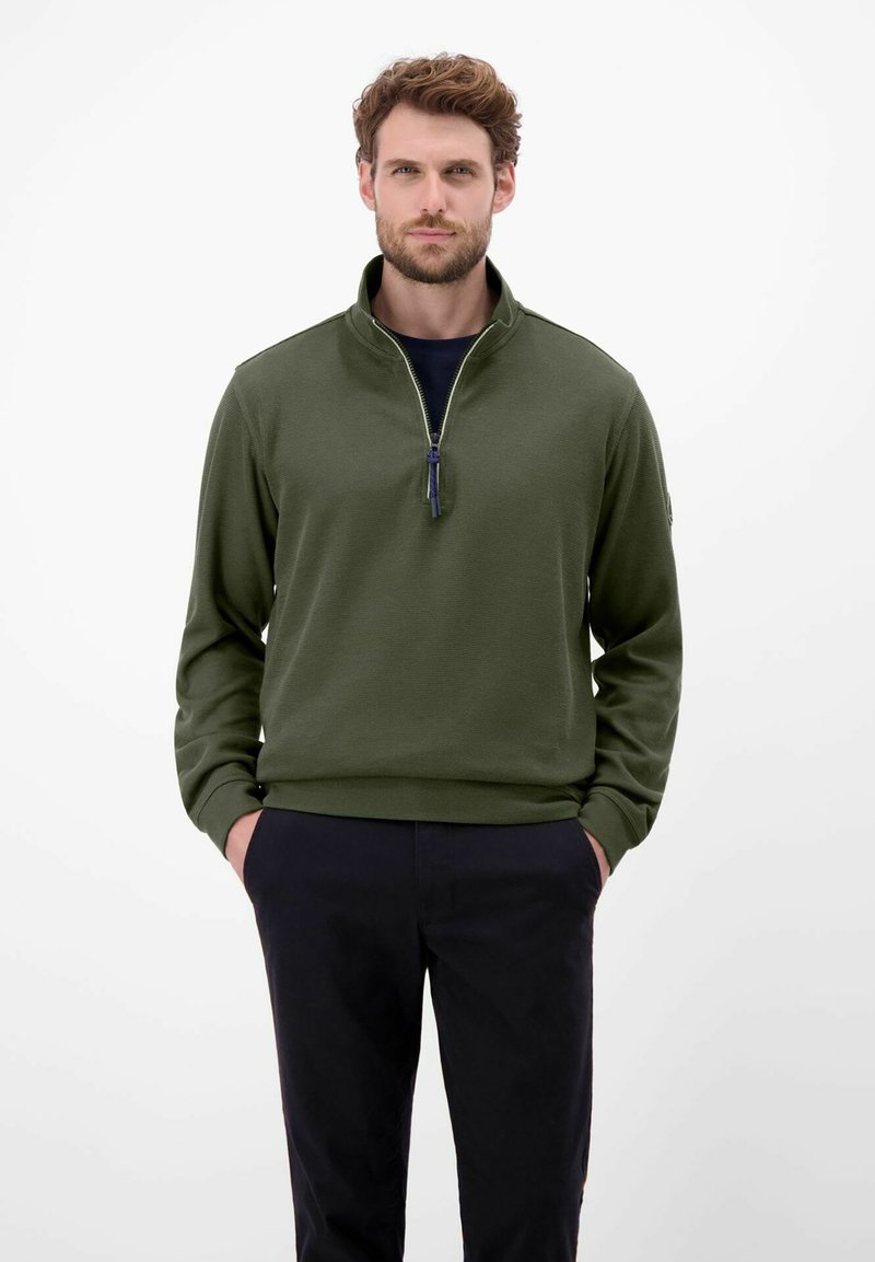 Mørkegrøn pullover med høj krave, halv lynlås og lange ærmer. Tekstureret stof, båret over en navy skjorte, parret med mørke bukser.