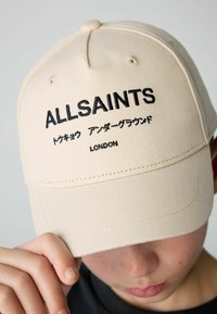 Beige szövetsapka, amelyen feketével hímzett "ALLSAINTS" és japán szöveg található. Kerek koronával és ívelt napellenzővel; minimalista dizájn.