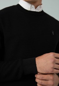 Hombre con un suéter negro texturizado sobre una camisa blanca de cuello, ajustando el puño del suéter con su otra mano.