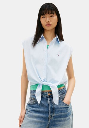 Giovane donna con capelli neri lisci indossa una camicia azzurra senza maniche, abbottonata e annodata in vita, sopra una maglietta verde e jeans blu a vita alta.