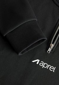 Black zip-up hoodie with ribbed cuffs, featuring a silver zipper and white logo "aprel" on the chest. Smooth cotton material, minimal design.