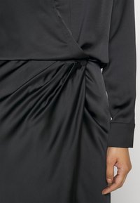 Robe portefeuille noire à manches longues avec tissu froncé et détail d'un bouton couvert à la taille, montrant le bras droit et la main.