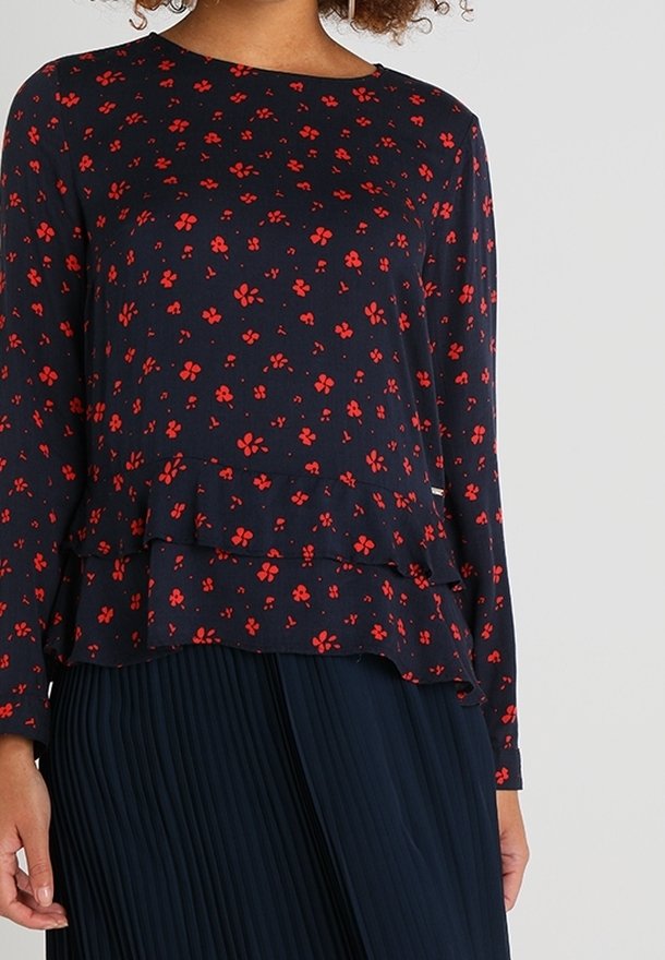 Femme portant un chemisier bleu marine à manches longues avec un petit imprimé floral rouge et un ourlet à volants superposés, associé à une jupe plissée bleu marine.