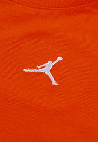 Orange stof med et hvidt broderet Jumpman-logo. Teksturen er glat med en ribbet kravedetalje.