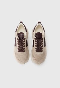 Baskets en suede beige avec accents marron foncé, présentant un bout arrondi, des détails cousus et un design à lacets. Texture lisse et semelle robuste.
