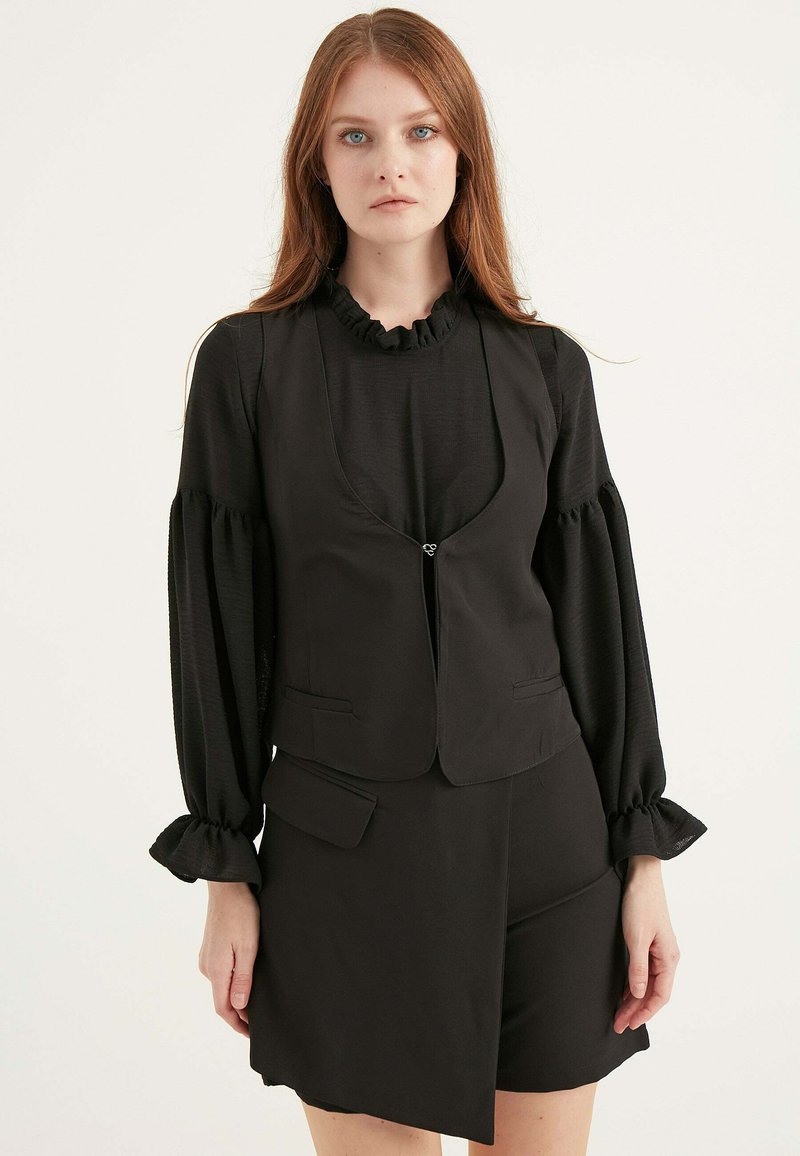 Svart blus med volangkrage och långa puffärmar, lager på lager under en figurnära väst med lätt rundad hals, med frampockets.