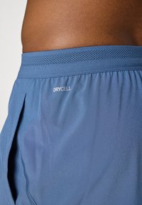 Pantaloni corti sportivi da uomo in tessuto blu, con una vita testurizzata e un piccolo logo DRYCELL. Il materiale sembra leggero e traspirante.