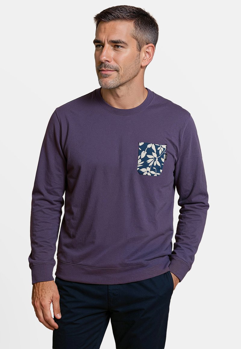 Paarse lange mouwenshirt met een marineblauw en witte bloemenzakdetail, ronde halslijn en geribbelde manchetten, gecombineerd met donkere broek.