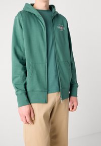 Felpa verde con zip realizzata in tessuto morbido, con tasca frontale, cappuccio regolabile con cordino e logo sul lato sinistro del petto. Abbinata a pantaloni corti beige.