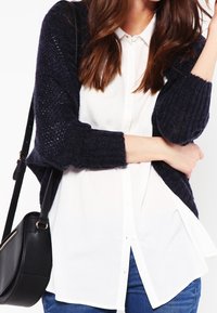 Námořnický pletený cardigan s otevřenými texturovanými rukávy, nošený přes bílou košili. Kombinováno s modrými džínami a černou kabelkou přes rameno.