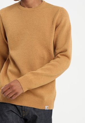 Stickad tröja i gyllenbrun, rund hals, ribbade ärmslut och fåll, med en liten logotagg på nederkanten. Strukturerad tyg.