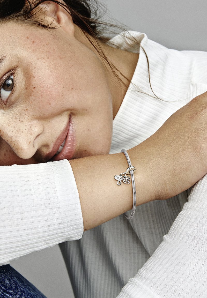 Strieborný náramok s príveskami v tvare srdca a kruhu, nosený na zápästí. Pozadie zobrazuje textúrovanú bielu košeľu.